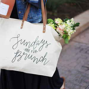 cropped image of a person carrying a canvas tote with leather handles. text on tote says "sundays are for brunch". There is a bouquet of flowers in the tote and the image was taken outdoors.