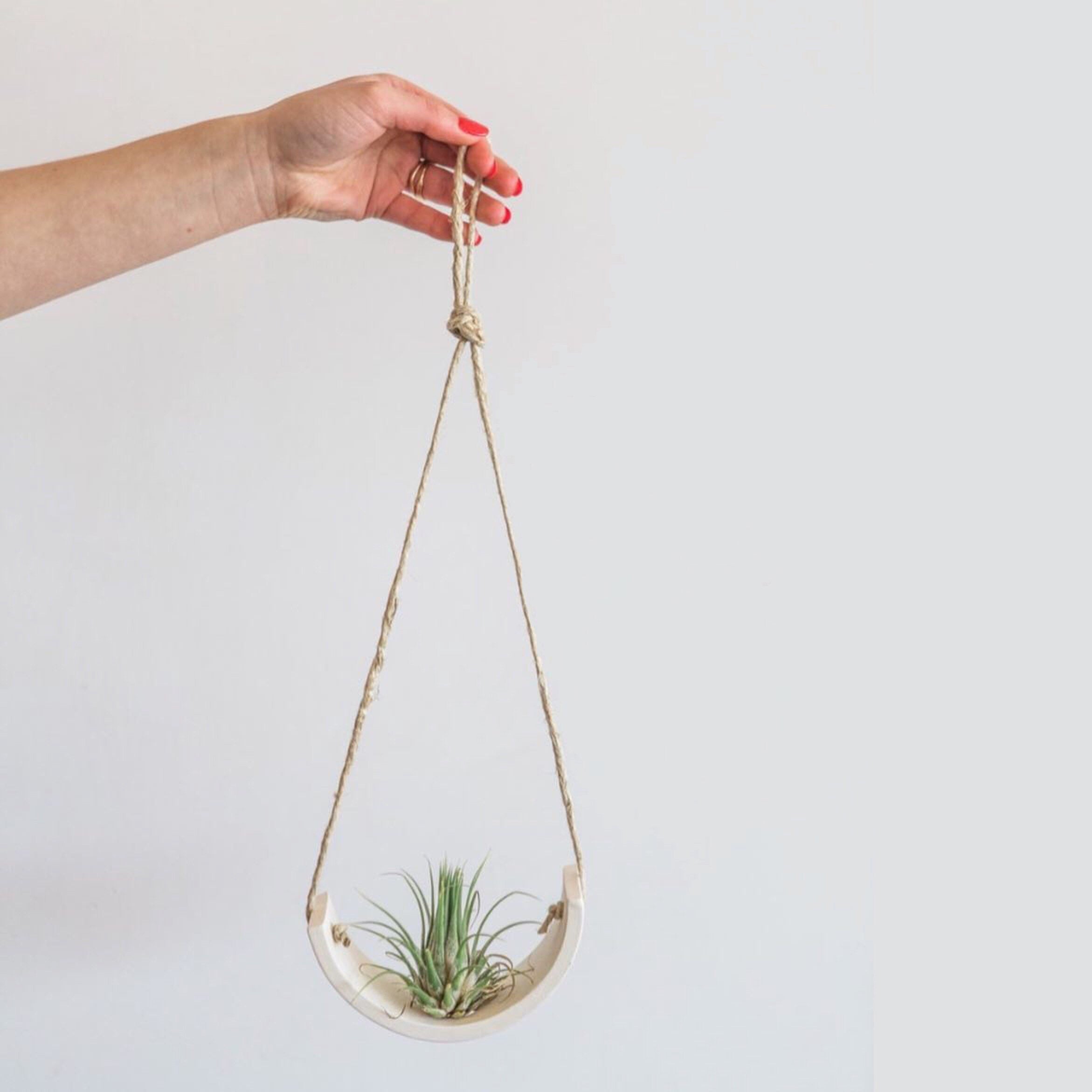 A white person’s hand holds up a white ceramic air plant hanger by a hemp cord.