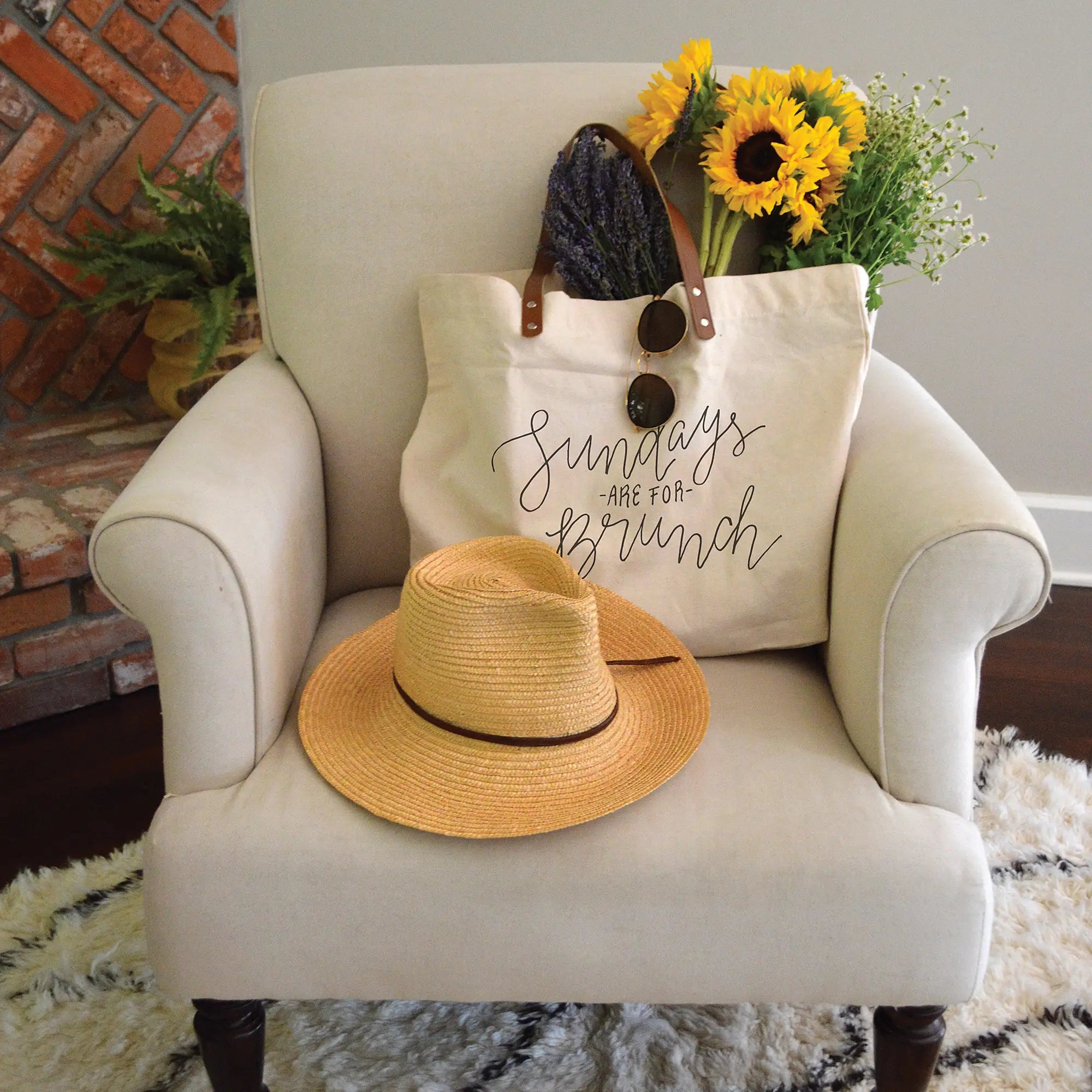 Indoor image of canvas totebag thay says "Sundays are for brunch". The tote is sitting on a white chair with a woman's hat and is filled with bouquets of flowers.