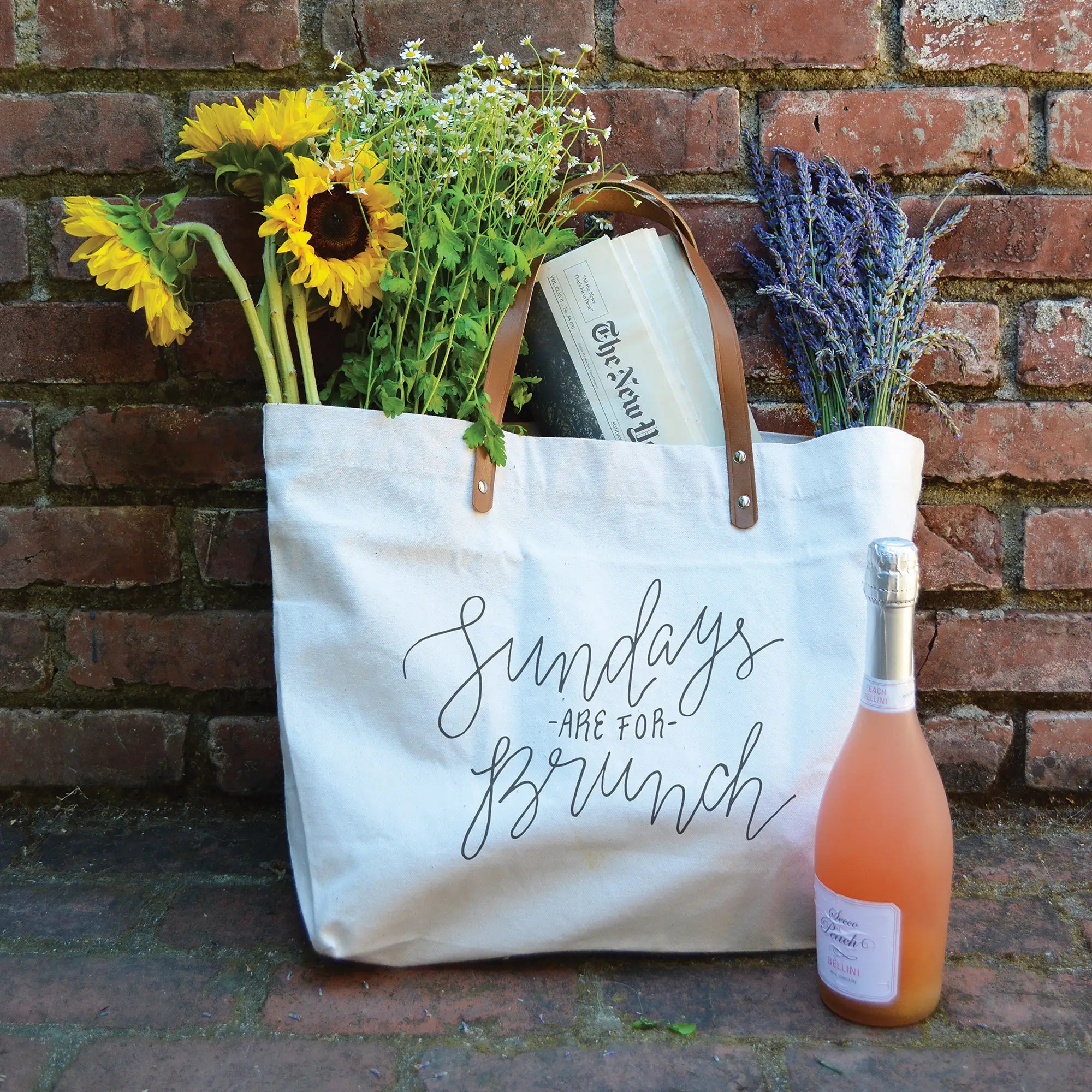 Tote bag with text "sundays are for brunch" against a brick wall. A bottle of sparkling wine is next to the bag and several bouquets of flowers are in the tote.